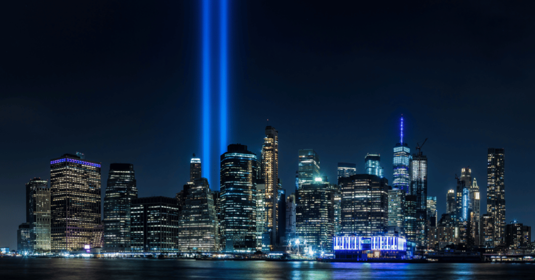 NYC landscape at night with two side-by-side spot lights shining their lights in the sky in remembrance of 9/11